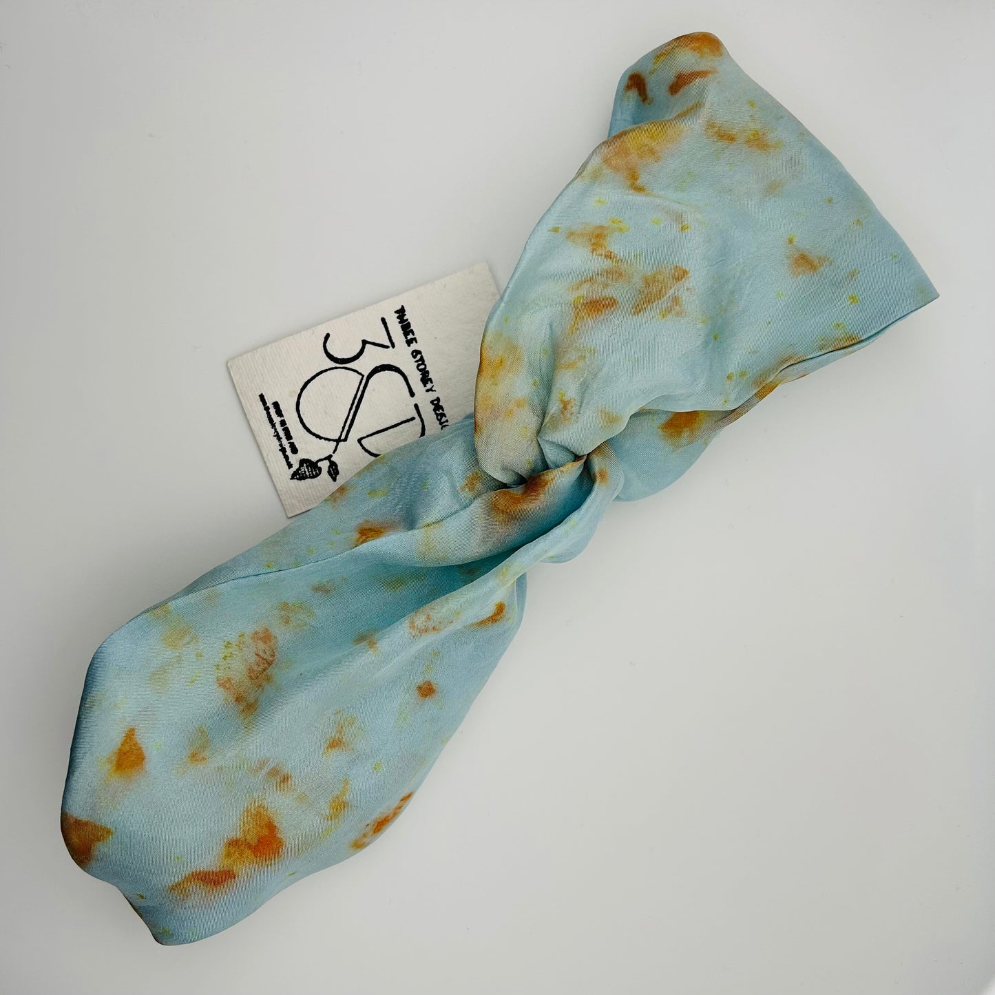 Top view of a Pale indigo silk turban headband with elastic to secure over head which has been naturally dyed with orange cosmos flowers